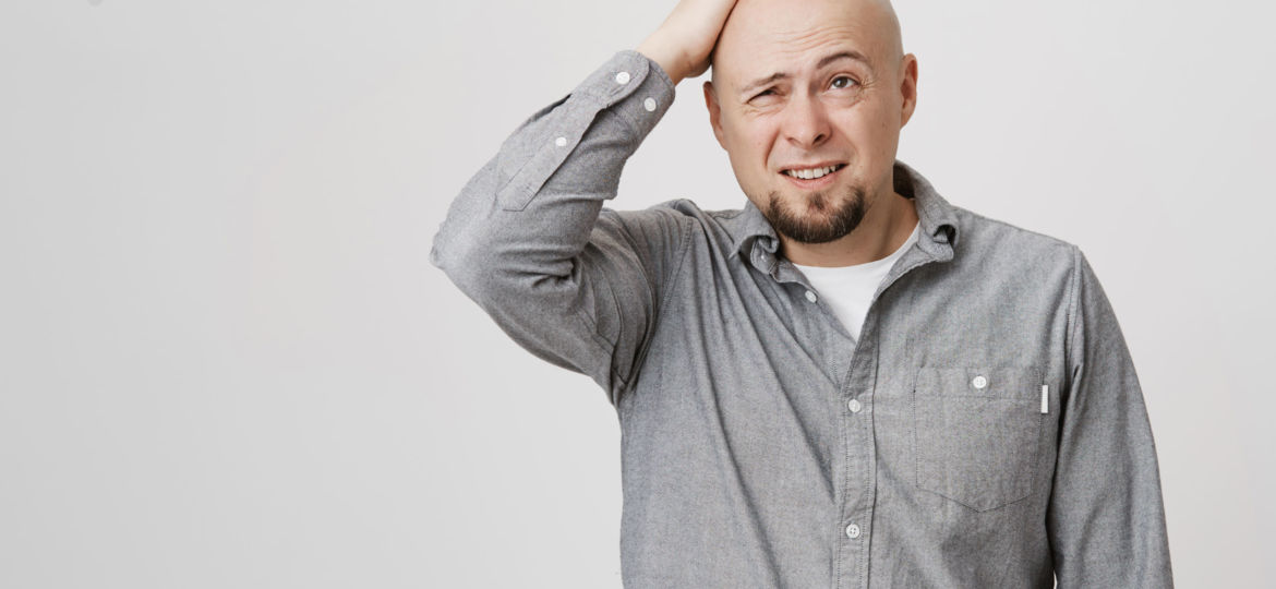 Picture of tired confused and questioned man holding his hand on head expressing ache after hit over white background. Handsome bearded guy thinks it is his turn to wash dishes.